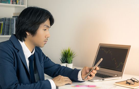 Young Asian Forex Trader or Investor or Businessman in Suit Looking Smartphone and Trading Forex or Stock Chart by Laptop in Trader Room in Vintage Tone