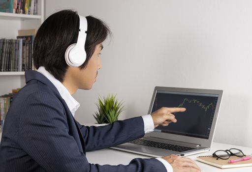 Young Asian Forex Trader or Investor or Businessman in Suit Trading and Pointing Forex or Stock Chart in Laptop Monitor in Trader Room