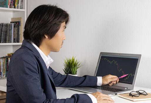 Young Asian Forex Trader or Investor or Businessman in Suit Trading and Pointing Forex or Stock Chart on Laptop Screen in Trader Room