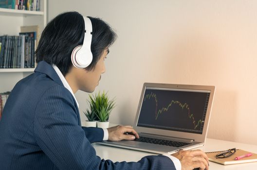 Young Asian Forex Trader or Investor or Businessman in Suit Wear Headphone and Trading Forex or Stock in Laptop Monitor in Trader Room in Vintage Tone