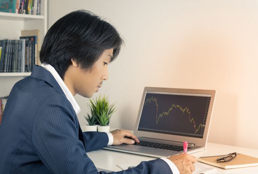 Young Asian Forex Trader or Investor or Businessman in Suit Writing Financial Report and Trading Forex or Stock in Laptop Monitor in Trader Room in Vintage Tone