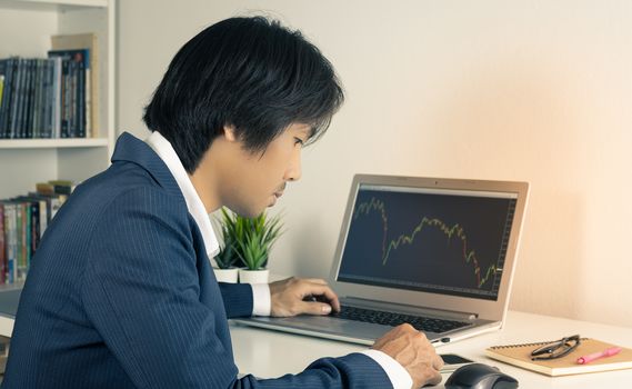 Young Asian Forex Trader or Investor or Businessman in Suit Touching Smartphone and Trading Forex or Stock in Laptop Monitor in Trader Room in Vintage Tone