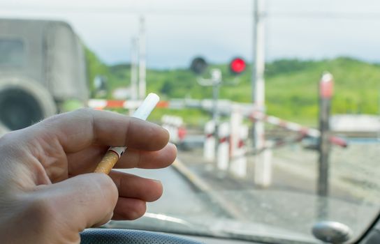 View of the driver hand with a cigarette on the steering wheel of the car, which stopped before a closed railway crossing at a red light