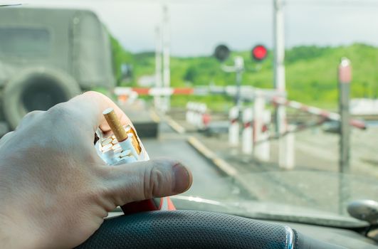 View of the driver hand with a pack of cigarettes on the steering wheel of the car, which stopped before a closed railway crossing at a red light