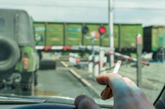View of the driver hand with a cigarette on the steering wheel of the car, which stopped before a closed railway crossing at a red light