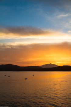 Sunrise on the Sardinian sea coast with intense orange color seen from the sea with five fishing boats on flat water