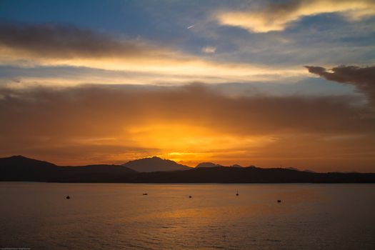 Sunrise on the Sardinian sea coast with intense orange color seen from the sea with five fishing boats on flat water