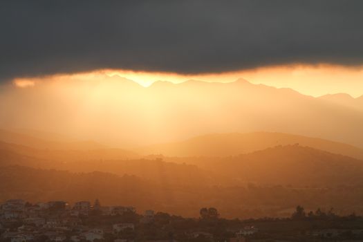 Breathtaking sunset after the storm. The orange sun's rays slip under a big black cloud illuminating the mountains and the hills with the houses of a small village