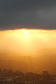 Breathtaking sunset after the storm. The orange sun's rays slip under a big black cloud illuminating the mountains and the hills with the houses of a small village