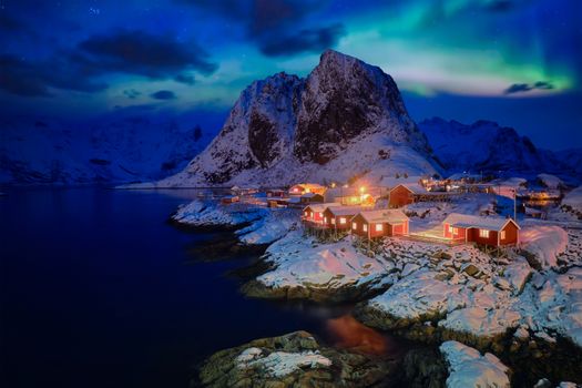 Famous tourist attraction Hamnoy fishing village on Lofoten Islands, Norway with red rorbu houses in winter snow illuminated in the evening with Aurora Borealis