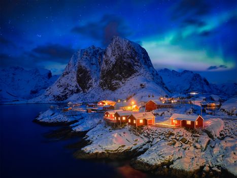 Famous tourist attraction Hamnoy fishing village on Lofoten Islands, Norway with red rorbu houses in winter snow illuminated in the evening with Aurora Borealis