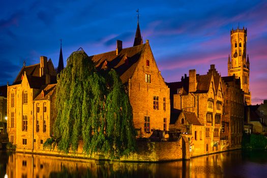 Famous view of Bruges tourist landmark attraction - Rozenhoedkaai canal with Belfry and old houses along canal with tree in the night. Brugge, Belgium