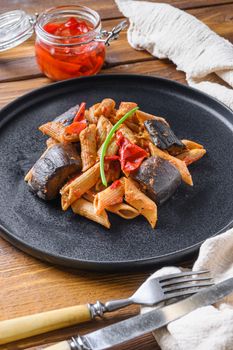 Aubergine penne eggplant pasta, pepper tomatoe sauce, on black plate over wooden table served side view close up.