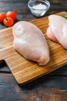 Raw chicken breasts and spices on wooden cutting board on old dark table side view close up.