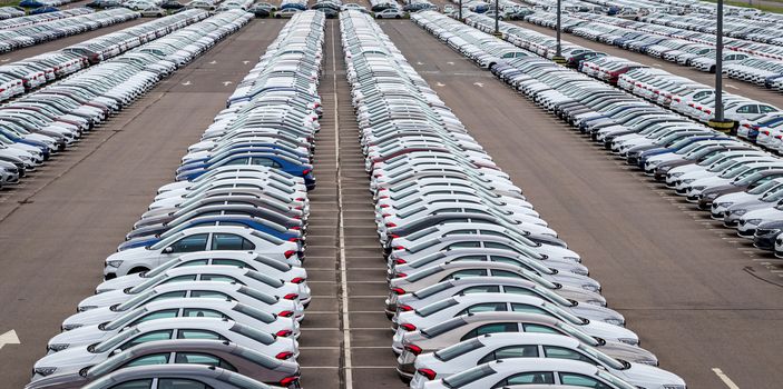 Rows of a new cars parked in a distribution center on a car factory on a cloudy day. Top view to the parking in the open air.