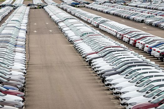 Rows of a new cars parked in a distribution center on a car factory on a cloudy day. Top view to the parking in the open air.