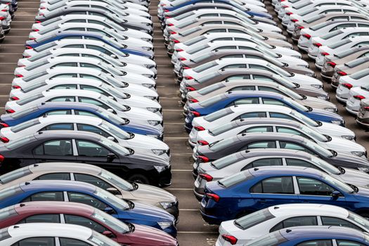 Rows of a new cars parked in a distribution center on a car factory on a cloudy day. Top view to the parking in the open air.