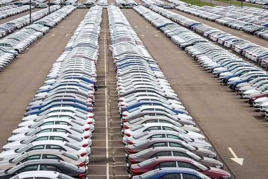 Rows of a new cars parked in a distribution center on a car factory on a cloudy day. Top view to the parking in the open air.