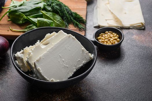 Traditional feta cheese in black bowl side view in front of ingredients.