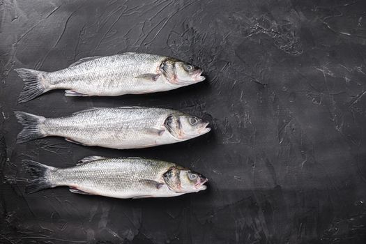 Set of whole raw sea bass with spices and herbs ingredients on chopping board over black stone table top view space for text