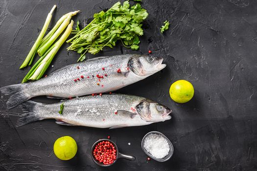 Set of whole raw seabass with spices and herbs ingredients on chopping board over black stone table top view space for text
