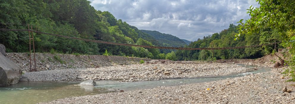 Suspension bridge over the mountain river at the summer cloudy day