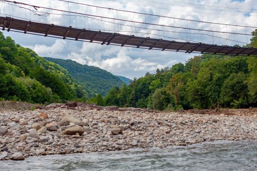 Suspension bridge over the mountain river at the summer cloudy day