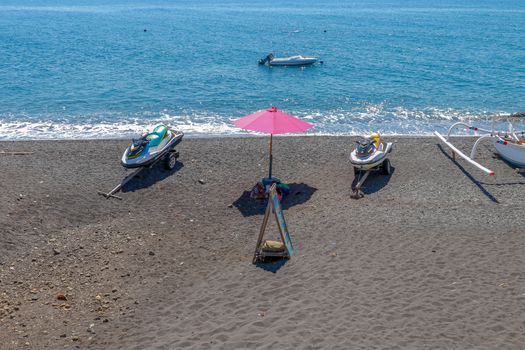 Jetski Seadoo parked on the beach. Black volcanic sand beach. In the background of the sea and moored boat. Red parasol.