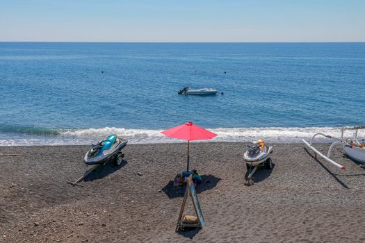 Jetski Seadoo parked on the beach. Black volcanic sand beach. In the background of the sea and moored boat. Red parasol.