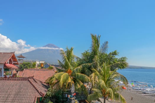 Tropical paradise with palm trees and majestic volcano in the background. White clouds around a volcano crater. View from above the roofs of the house.