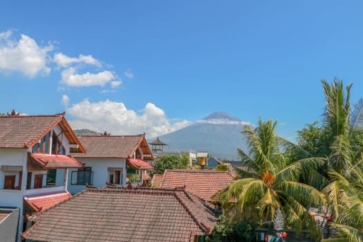 Tropical paradise with palm trees and majestic volcano in the background. White clouds around a volcano crater. View from above the roofs of the house.