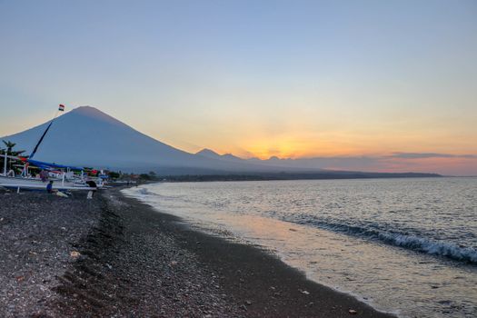 Sunset on the beach on a tropical island. Orange colored sky and clouds. Big majestic volcano on the horizon. Quiet sea and romantic atmosphere.