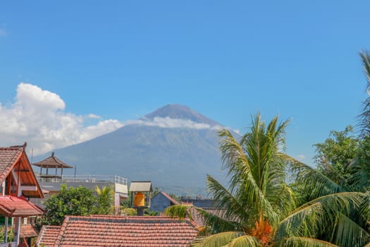 Tropical paradise with palm trees and majestic volcano in the background. White clouds around a volcano crater. View from above the roofs of the house.