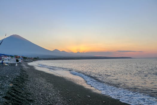 Sunset on the beach on a tropical island. Orange colored sky and clouds. Big majestic volcano on the horizon. Quiet sea and romantic atmosphere.