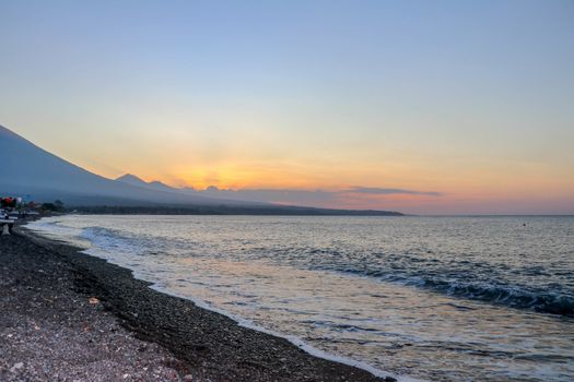 Sunset on the beach on a tropical island. Orange colored sky and clouds. Big majestic volcano on the horizon. Quiet sea and romantic atmosphere.