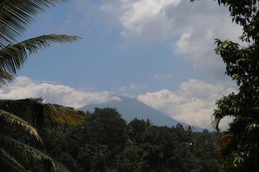 View through the trees in the foreground to the active Gunung Agung volcano from the opposite hill on Bali Island in Indonesia. Azure blue sky with dense white clouds on the sides of the mountain.