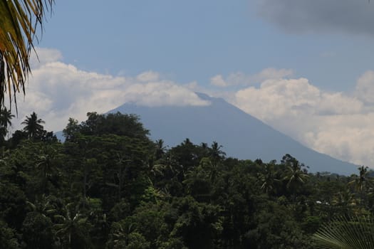View through the trees in the foreground to the active Gunung Agung volcano from the opposite hill on Bali Island in Indonesia. Azure blue sky with dense white clouds on the sides of the mountain.