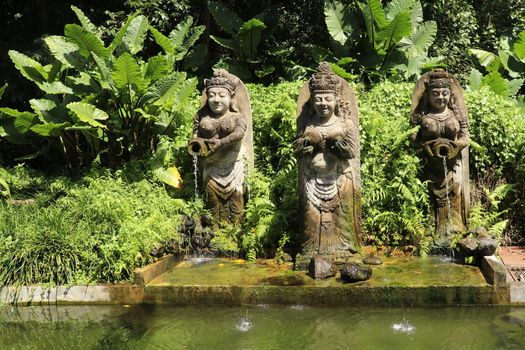 Three Statues in Bali Indonesia of a fountain with jungle in the background. Balinese statuette of a girl with a pot from which water flows. Pool decoration.