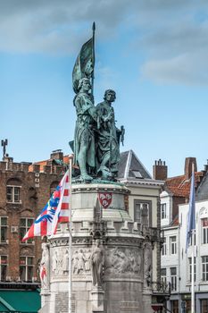 Bruges, Flanders, Belgium -  June 15, 2019: Green bronze statue of Jan Breydel and Pieter De Coninck on Mark Square against light blue sky. Gables of north corner and flag in back.