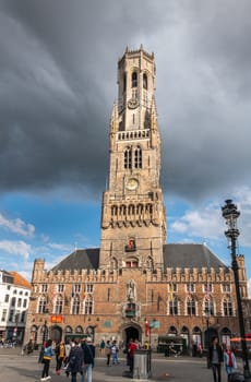 Bruges, Flanders, Belgium -  June 15, 2019: Frontal view on the brown stone Halletoren building, named Belfort or Belfry, and clock tower against dark gray cloudscape. People on the Markt, square.