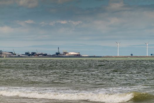 Knokke-Heist, Flanders, Belgium -  June 16, 2019: Knokke-Zoute part of town. View from sea edge on LNG sea terminal and Wind turbines of Port of Zeebrugge. People on sand. Greenish Nord Sea,