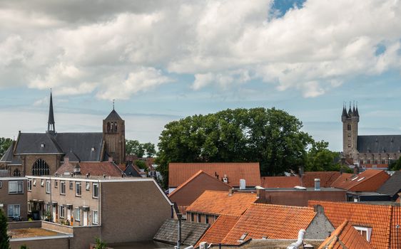 Sluis, the Netherlands -  June 16, 2019: View over red roofs at two towers of the town under blue sky with white cloudscape, Green foliage of tree in center.