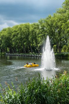 Sluis, the Netherlands -  June 16, 2019: People in yellow pedal boat pass the fountain in the center of town, located in canal to Dammy, Belgium. Green curtains of reed and trees. Cloudscape.