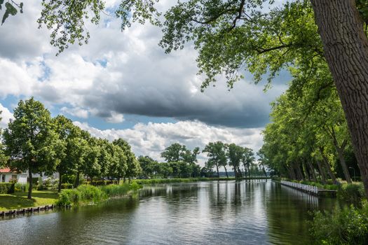 Sluis, the Netherlands -  June 16, 2019: Reflecting water of canal to Damme in Belgium, with curtains of green trees on sides, under a storm approaching cloudscape.