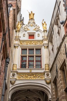 Bruges, Flanders, Belgium -  June 16, 2019: Bridge is room links city hall and Brugse Vrije building over Blinde Ezelstraat. Silver sky between brown buildings. Golden statues