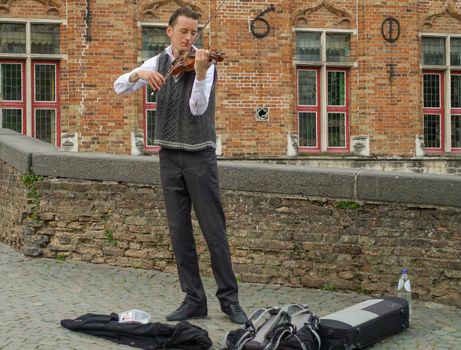 Bruges, Flanders, Belgium -  June 16, 2019: Young man plays violin as beggar for money on bridge over canal at Blinde Ezelstraat. Red brick building in back.