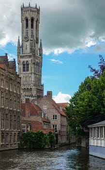 Bruges, Flanders, Belgium -  June 16, 2019: Shot of historic Brugge with Belfry across canal from Huidevettersplein. Brown stone buildings, green foliage and heavy cloudscape.