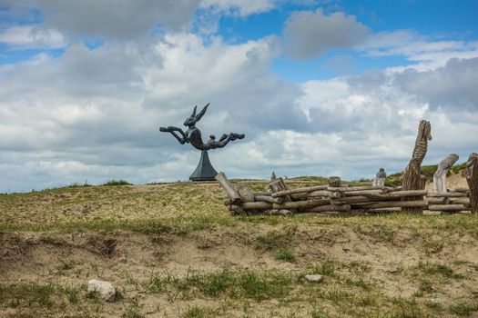 Knokke-Heist, Flanders, Belgium -  June 16, 2019: Knokke-Zoute part of town. Jumping hare statue on brown-green dune behind pile of wood. Captured in  blue and white cloudscape.