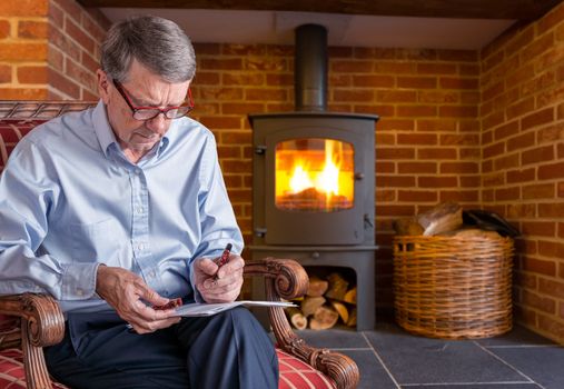 Senior caucasian adult checking a document with fountain pen in hand while seated. He is sitting by wood burning furnace
