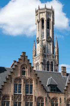 Bruges, Flanders, Belgium -  June 17, 2019: Gray stone tower of Belfry with clock peeps over brown brick step gable under blue sky with whtie cloud.